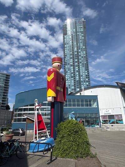 This is a landscape-oriented, vertical photograph taken on a sunny day with scattered white clouds against a bright blue sky. The primary subject is a large, wooden statue of a bellhop dressed in a red uniform with yellow accents and a matching cap. The bellhop is positioned in the foreground, facing left, with a slightly smiling expression.
Behind the statue, a modern, blue building with large windows and a curved roofline is visible. To the left of the building, a very tall, multi-story residential building with many windows dominates the background. A wooden boardwalk runs in front of the blue building. To the right of the bellhop, a blue kayak is secured in a bike rack, with a red bu oy-like structure mounted nearby. Bicycles are parked in front of the kayak.
In the distance, other buildings and trees can be seen. The overall scene suggests an urban waterfront or a public recreational area. The lighting and shadows indicate it is likely midday. There is no discernible activity taking place beyond people passing by or using the facilities.
Visible text includes "Fraser River" on a sign above the entrance to the blue building. The phrase "play. explore. discover!" is also visible on the blue building's exterior. The location context is New Westminster, Canada.