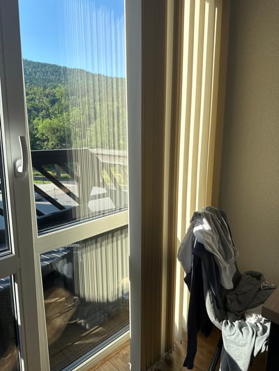 This media file shows an interior view of a room with a large window overlooking a natural landscape. The room appears to be a bedroom or living space, with blinds drawn in front of the window. A pile of clothes, consisting of various gray and white garments, is visible on top of the blinds. The scene suggests a domestic setting, likely in a mountainous or hilly area given the view outside.
The window offers a glimpse of a lush green hillside covered in trees, under a clear blue sky. The sunlight streaming through the blinds creates a pattern of light and shadow on the wall and the pile of clothes. The time of day appears to be morning or afternoon, indicated by the bright sunlight and the angle of the shadows. The weather seems to be pleasant and clear, with no signs of rain or clouds.
The location is identified as Szklarska Poręba, Poland, a town known for its scenic beauty and mountainous terrain. This context aligns with the visual cues of a forested hillside visible through the window. The overall mood is serene and quiet, with a sense of domestic comfort juxtaposed with the natural beauty of the surroundings. There is no visible text in the image.