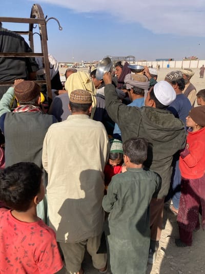 This image depicts a densely crowded outdoor scene in Chaman City Tehsil, Pakistan, under a clear sky. A large group of individuals, predominantly adult males and several children, are gathered closely together, facing towards the left side of the frame where a dark, metallic structure, possibly part of a truck or makeshift stand, is visible. Many individuals wear traditional Pakistani attire, including shalwar kameez and various types of kufi caps and turbans.
Multiple people are holding empty, shiny metal bowls or plates aloft, suggesting they are awaiting or attempting to receive a distributed item, likely food or water. One man in the center-right, wearing a dark green jacket and a white kufi, has his right arm raised, holding a metal bowl. Another man to his left also holds a bowl above his head. Children are interspersed within the crowd; one young boy in a red shirt is in the foreground, facing away from the camera, and another in a dark green shirt stands behind him. Long shadows cast by the individuals indicate a bright, sunny day.
The background reveals an open, dusty, unpaved area with various distant structures, including a white boundary wall, what appear to be container-like structures, and utility poles, consistent with a border region or camp environment. The overall scene conveys a sense of anticipation and urgency among the gathered people.