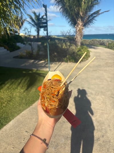 A person holds a wooden container filled with noodles, vegetables, and a yellow sauce. Two chopsticks are placed on top. A red packet is visible next to the container. The background features a tropical landscape with palm trees, grass, and a walkway. The ocean is visible in the distance. The person's shadow is cast on the ground, and a beaded bracelet is seen on their wrist.