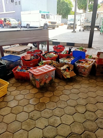 The scene depicts an outdoor setting in Awoyaya, Nigeria, characterized by various containers filled with food items. The containers include red and yellow plastic baskets, along with transparent and blue plastic storage boxes, as well as cardboard boxes, all brimming with packaged food. These containers are resting on a ground paved with hexagonal concrete tiles.
In the background, several commercial vehicles or trucks are parked. A group of people are seen near one of these vehicles, while the remaining vehicles are unoccupied. A pallet and a wooden structure (possibly a table or a bench) are visible in the background as well.