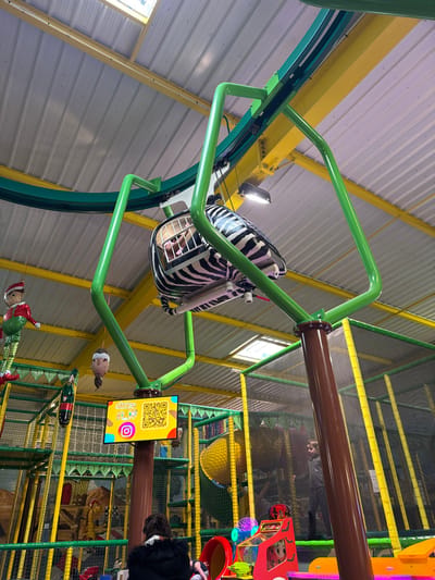 This image depicts the interior of Kid's Paradis, an indoor children's play park located in Bénouville, France. The scene is brightly lit, featuring a high, corrugated metal ceiling with exposed yellow structural beams.
Dominating the upper portion of the frame is a green overhead monorail system, supported by green and brown-painted structures. A single zebra-striped capsule, or gondola, is suspended from this track, moving from left to right. Inside the capsule, a child wearing a pink garment is visible, facing forward.
Below the monorail, a tall, brown cylindrical column, designed to resemble a tree trunk, rises from the floor to support the monorail track. A flat-screen monitor is mounted on this column, displaying the "Kid's Paradis" logo, multiple QR codes, and social media icons (Instagram, Facebook, YouTube).
Various festive decorations hang from the ceiling and overhead structures: a large red and green elf figure, a brown ornament, and a black and gold champagne bottle decoration.
The background shows a multi-level soft play structure constructed with yellow and green padded poles and green netting. A large yellow tunnel slide is visible within this structure.
In the foreground, at the bottom center, an individual with dark hair, wearing a black jacket with a fur-trimmed hood, is partially visible from behind, facing away from the camera. To the right, within the netted play structure, another individual in a dark jacket is standing. Portions of colorful arcade games or similar play equipment, predominantly red and purple, are visible on the ground level to the right.