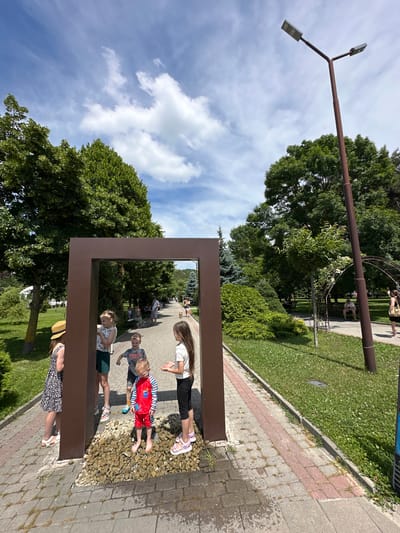 The image depicts a sunny day in a park, likely in Abrau-Dyurso, Russia, given the context. A large, brown, rectangular frame is the central focal point, positioned on a walkway paved with interlocking stones and bordered by a strip of pinkish stones. The frame stands on a bed of pebbles, through which water appears to be flowing from a grate.
Several children are gathered around this frame, interacting with it and each other. On the left, a young girl wearing a straw hat and a black and white patterned dress stands with her back to the camera. To her right, inside the frame, a girl in a white sleeveless top and dark shorts is visible, looking towards the right. In front of them, a boy in a red and blue striped swimsuit stands barefoot on the pebbles, looking down with a slightly distressed expression. To the right of the boy, a girl with long dark hair, wearing a white t-shirt and black leggings, looks towards the boy. Another boy, wearing a grey t-shirt with a graphic print, is partially visible behind the girl in the white top, appearing to gesture.
The park setting features lush green trees and manicured lawns with small white flowers. A paved path, similar to the one in the foreground, runs alongside the grassy area. In the background, people are seen relaxing on benches and walking. A tall, dark brown lamppost with two lights is prominently positioned to the right of the frame, casting a shadow on the path. An arched trellised structure covered in greenery is also visible in the background. The sky is blue with scattered white clouds.