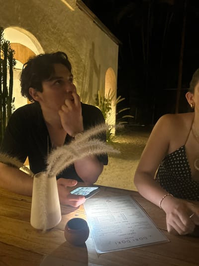The scene is an outdoor restaurant at night, located in the Cobano District of Costa Rica (9.635, -85.158). Two people, a man and a woman, are seated at a wooden table. The man is wearing a black shirt and is looking off to the side. He is holding a cell phone in his left hand and touching his face with his right. The woman is wearing a black and white polka-dot tank top and is looking down at the table. She is wearing a bracelet on her right wrist. There is a tall, white vase with white feather-like plants in it on the table. The menu, written in Spanish, sits on the table. There is a brown cup to the right of the vase. The scene is lit by warm, overhead lighting. The restaurant is surrounded by tall trees. The ground is covered with sand. The wall behind the man is white and has an arched opening. It is covered in shadows. There is a large cactus plant in the background on the left side of the frame.