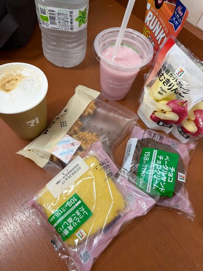 The image displays a selection of convenience store food items laid out on a wooden surface, likely a table. In the upper left corner, a portion of a black bag is visible. To its right, a clear plastic bottle of water with a white label containing Japanese text and a QR code is present. In the foreground, a white paper cup with the "SEVEN CAFE" logo holds a frothy, light brown beverage, possibly coffee or latte. Next to it, a clear plastic cup with a white lid and straw contains a pink smoothie, also with the "SEVEN CAFE SMOOTHIE" branding.
Several packaged food items are arranged around these drinks. To the right of the coffee cup, a beige and white striped wrapper encloses a long, narrow pastry. Further right, a bag of sliced apples is visible, with two whole apples and several apple slices displayed. Above the apples, an orange and white package labeled "SNACKY CHOCOLATE" is prominent.
Below the apples and smoothie, a dark brown, round pastry in a clear plastic wrapper is labeled "チョコレートクリーム" (Chocolate Cream) and "ベーグルサンド" (Bagel Sandwich). To the left of this, a yellow, rectangular cake-like item in a pink and clear wrapper is visible, with a green label that reads "たまご蒸しパン" (Steamed Bread with Egg). A white label with Japanese text, including a date and characters like "+YMK", is attached to this yellow pastry.
The overall scene suggests a casual meal or snack break, possibly at a convenience store or a seating area within one. The lighting is natural and even, and the image is taken from a slightly elevated, top-down perspective. The context of "Chuo, Japan" for the city location indicates a potential real-world setting where these items might be purchased or consumed.