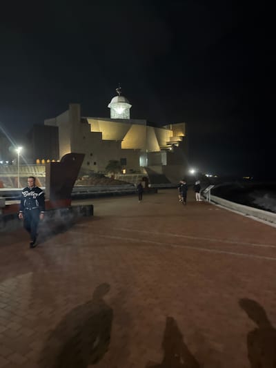 A nighttime exterior scene shows a paved walkway in front of a large, illuminated building in Las Palmas De Gran Canaria, Spain. The building, with its textured walls and geometric design, is lit by warm artificial lights, highlighting its architectural features, including a domed structure on top that also emits light. In the foreground, a man wearing a black jacket with white lettering and a white stripe on the sleeves is walking towards the left side of the frame. Several other individuals are visible further down the walkway, some walking and others standing. The walkway itself is made of reddish-brown bricks laid in a pattern, with white lines indicating lanes or boundaries. To the right of the walkway, the dark ocean is visible, with waves breaking against a concrete barrier. Shadows of people are cast on the walkway. The overall atmosphere is one of a public promenade at night, with the building serving as a prominent landmark.