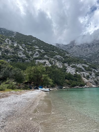 This image depicts a beach in Kumluca, Turkey. On the right, there is a pebble beach transitioning to clear, turquoise water. Adjacent to the beach, there's vegetation with trees, a small wooden structure, a blue trailer, and a white boat. Rising on the left is a steep, rocky mountain partially obscured by low-hanging clouds. The weather appears overcast.