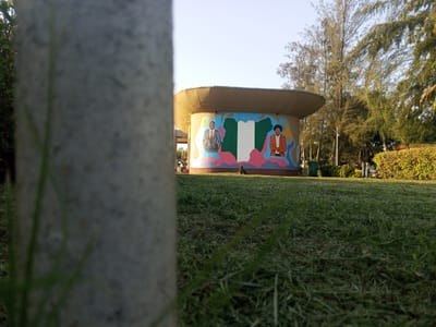 The image is taken from a low angle in a park in Tawpo, Nigeria. A blurred grey concrete pillar dominates the foreground. Beyond the pillar is a green lawn, and further back is a cylindrical building with a flat roof. The building has a mural depicting two people and the Nigerian flag. Trees and shrubs surround the building. The sky is light blue.
