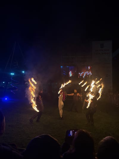 This nighttime image captures a fire performance during an outdoor festival in Elin Pelin, Bulgaria. In the foreground, the silhouetted heads of several spectators are visible, with one individual on the lower right filming the event horizontally with a smartphone, its screen showing a bright, blurry reflection of the flames.

The central focus is on four performers on a grassy area. From left to right: a performer in dark attire creates a vertical line of intense flames and smoke with a fire prop, facing right. To their right, a second performer, dressed in a light-colored, form-fitting suit with pink hair, holds a flaming hoop, facing towards the right. A third performer, with dark hair and wearing dark costume elements, stands center-right, manipulating several flaming hoops to create a broad fan-like display of fire. The fourth performer, positioned on the far right and also in dark clothing, mirrors this action with multiple flaming hoops, facing towards the left. The ground is green grass, illuminated by the fire and ambient blue stage lighting.

In the mid-ground on the left, a tall, slender structure with a glowing red and blue spiral design ascends, with blue spotlights at its base. In the background on the right, a large white banner is prominently displayed, featuring the Cyrillic text "ШОПСКИ ПРАЗНИК" (Shopska Feast) at the top, along with smaller sponsor logos like "FLYNET" and "NOBEL." Below the banner, a large screen exhibits abstract, vibrant light patterns. Scaffolding structures support the banner and screen. Beyond the performance area, behind a metal barrier, a dimly lit crowd of additional spectators can be discerned. The sky above is dark, indicative of nighttime.
