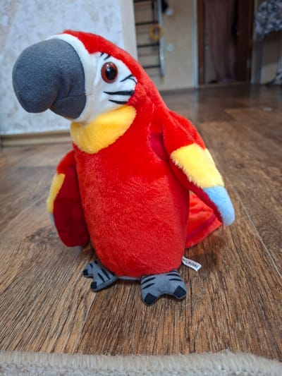 A plush toy parrot with red, yellow, blue, and gray coloring is sitting on a wooden floor. The parrot is positioned facing left and is slightly angled towards the viewer. Its beak is gray, and its eyes are red with black pupils. Yellow and white feathers surround its neck, and red and yellow feathers adorn its wings. The parrot's feet are gray with black stripes. A small white tag that reads "MADE IN CHINA" is attached to its body. The background shows a wall with a light-colored texture, a portion of a door frame, and some blurred objects. The floor is made of wood planks with visible grain. This scene is located in Gorkovskiy, Russia.