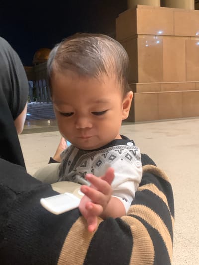 The image features a close-up, slightly tilted view of a baby or young toddler being held by an adult. The baby, positioned prominently in the left half of the frame, has dark, slicked-back hair and a soft, downward gaze, suggesting curiosity or focus on something below. Their right ear is visible.

The child is wearing a white short-sleeved top adorned with a black and white geometric pattern, possibly a traditional or tribal design, particularly visible on the front and shoulders. Their left hand is gently touching a small, white rectangular tag or label, which appears to be attached to the adult's clothing. The adult, whose face is not visible, is wearing a garment with prominent black and tan horizontal stripes on the right side, likely a sweater or long-sleeved top. A plain black fabric is also visible in the lower left, possibly part of the adult's attire.

The background is softly lit, indicating an indoor setting or a covered outdoor area at night or in the evening. Light-colored, possibly tiled or stone, architectural elements are visible in the upper background, with reflections from artificial light sources creating soft glows. A darker, less defined area is present in the lower background. The scene conveys a quiet, tender moment between the child and the unseen adult. No visible text is present in the image. The provided location context is Samarinda, Indonesia.