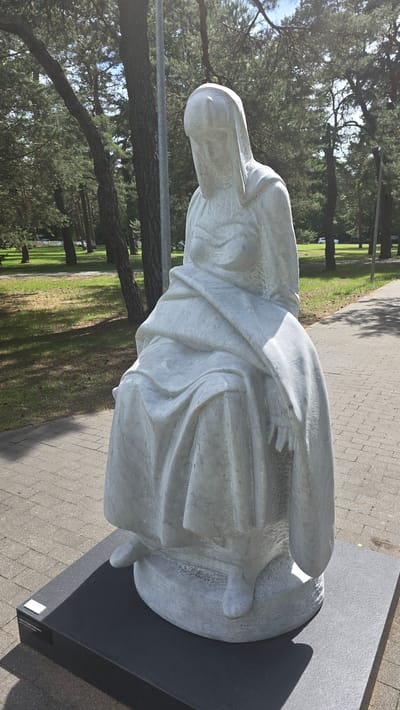 This media file showcases a white marble sculpture of a robed figure, seated and looking down. The sculpture is detailed with flowing drapery that suggests fabric. The figure's face is partially obscured by its hood, but the impression is of a contemplative or sorrowful expression. The sculpture is placed on a dark rectangular base, which sits on a paved walkway.
The setting appears to be an outdoor public space, possibly a park or garden, with a pathway made of interlocking paving stones. Lush green grass and mature trees with dark bark and green foliage surround the walkway, indicating a natural or landscaped environment. The lighting suggests it is daytime, with sunlight casting shadows from the trees onto the pathway. The overall atmosphere conveyed by the sculpture and its surroundings is one of solemnity and tranquility.
There is a small white plaque visible on the dark base of the sculpture, which likely contains information about the artwork, such as the artist's name and the title of the piece. Although the text on the plaque is too small to read clearly in the image, its presence indicates this is a public display. The location is identified as Konstancin-Jeziorna, Poland.