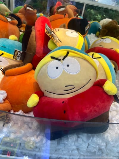 A collection of plush toys, primarily characters from the animated television series South Park, are displayed inside a claw machine. The most prominent toy in the foreground is Eric Cartman, recognizable by his red jacket, yellow hat, and distinctive facial features. Other plush toys visible include Kenny McCormick, identifiable by his orange parka and hood, and possibly Kyle Broflovski and Stan Marsh due to their yellow hats and blue/green accents. The toys are nestled among white packing peanuts. The claw machine's glass front reflects some of the surrounding environment, including a hint of greenery, suggesting an outdoor or semi-outdoor location. A small digital display on the machine shows "U" and "Credits 0," indicating it is not currently in use. The toys appear to be new, with at least one plush having a "South Park" tag attached. The scene suggests a retail or amusement setting, possibly a shopping mall or arcade. The question about its location in Baunatal, Germany, cannot be answered from the image itself.
