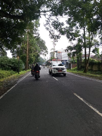 A forward-facing street-level view depicts a paved, two-lane road under an overcast sky. Lush green foliage from tall trees and dense undergrowth lines both sides of the road, with large tree branches overhanging the roadway. In the left lane, two dark motorcycles are visible moving away from the camera; the lead rider wears a dark helmet. In the opposite, right lane, a white Chevrolet Colorado-style utility vehicle, fitted with red and blue emergency lights on its roof and displaying a red front license plate with "F 9032 HD," is approaching. In the mid-distance on the right, behind a low wall partially obscured by vegetation, are several multi-story buildings with white and grey facades and visible commercial signage. A streetlight pole is present on the left side of the road in the mid-ground. The scene portrays active road traffic within a verdant, semi-urban setting.