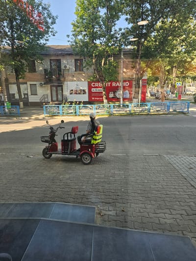 A person wearing a high-visibility vest sits on a red three-wheeled scooter parked on a paved sidewalk next to a street. The scooter has a basket in the front and a storage compartment behind the seat. In the background, a building with a brick facade has a large advertisement for "CREDIT RAPID" visible on its exterior. Trees with green foliage line the street, and the sky is clear. A blue metal fence separates the sidewalk from the street in some areas.