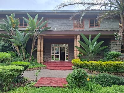The image shows the front entrance of a building located at latitude -0.754 and longitude 36.437 in the city of Karagita, Kenya. The building has a stone facade and is two stories tall. The entrance is framed by two large banana plants and a  brick walkway. The entrance is a double door with glass panels that lead into the interior. There are several windows on the upper story of the building. The building is surrounded by manicured bushes and landscaping. There are no people visible in the image.