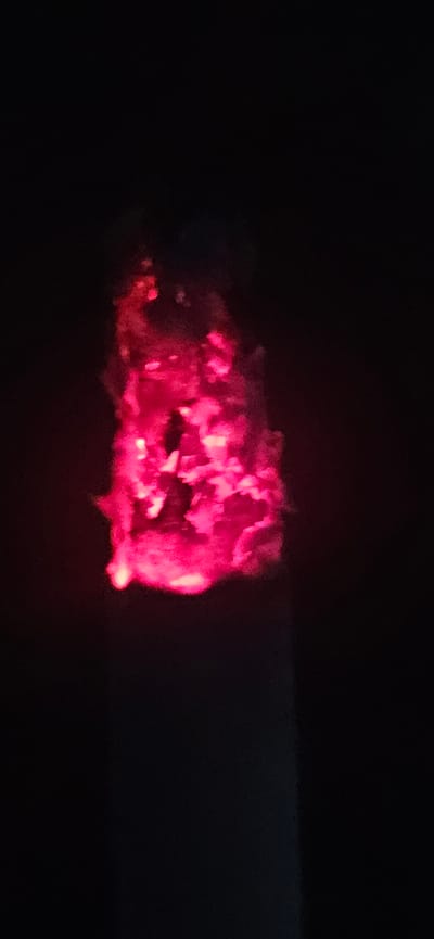The image is a close-up, low-light photograph of a burning object, possibly embers or coals, glowing intensely with a vibrant pink and red hue against a predominantly black background. The object appears to be irregular in shape and is surrounded by wisps of smoke or glowing particles. There is a vertical shaft of light towards the bottom right of the image. The overall impression is one of intense heat and combustion in a dark environment. There are no discernible features indicating a specific location in Momin Prohod, Bulgaria, or any other identifiable geographical markers.