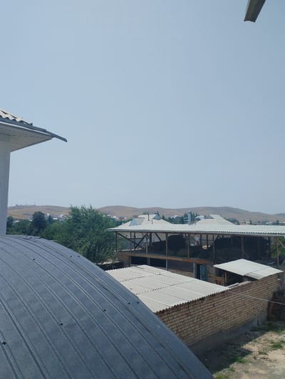The image captures a view of the city of Mady, Kyrgyzstan, under a clear sky. In the foreground, a dark, corrugated metal roof dominates the left side of the frame, curving upwards. To the right, a low-rise brick building with a corrugated roof is visible, its walls constructed from light-colored bricks. Adjacent to this building is an open-air structure with a white corrugated roof, supported by wooden beams, and appears to be storing hay or similar agricultural material. Beyond these structures, a dense green area of trees partially obscures a cluster of houses with light-colored, sloped roofs, nestled against rolling, barren hills in the background. The overall scene suggests a rural or semi-rural setting within Mady.