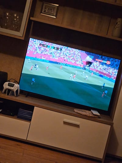 A television screen displays a soccer match with a score of 0-1. The clock shows 46:21. The teams represented appear to be Germany (GER) and Ivory Coast (CIV), indicated by national flags next to their scores. The broadcast includes sponsor advertisements for "ADIPREDICTSTREET.COM" and "ADIPREDICTSTREE." The television is situated on a wooden media console in what appears to be a living room environment. A white video game controller is placed on a charging dock to the left of the television. The media console has light-colored drawers and wooden accents. Behind the television, a wooden shelf holds decorative items, including a framed picture and a vase. The overall setting suggests a home entertainment area in Novi Iskar, Bulgaria, where the broadcast is being watched.