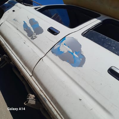 The image shows a white, heavily damaged car door with significant paint chipping and exposed metal. The paint loss reveals a light blue underlayer. The door handle is chrome. The car is situated in an outdoor environment, possibly a junkyard or repair shop. The location is identified as Khan Yunis, Unknown. The lower portion of the car is visible, with a corroded metal support structure. The image was taken with a Galaxy A14 phone, as indicated by the watermark.