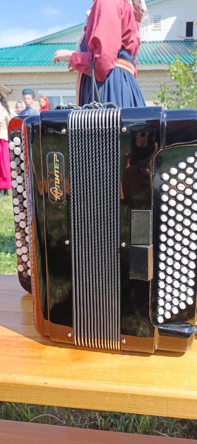 A black and silver "Jupiter" button accordion sits on a wooden surface in an outdoor setting. The accordion's bellows are visible. A person wearing traditional red and blue clothing stands in the background, along with a group of individuals. The location is likely Кемуль, Russia, given the logo on the accordion. The accordion is on a wooden surface. Green grass is visible around the wooden surface.