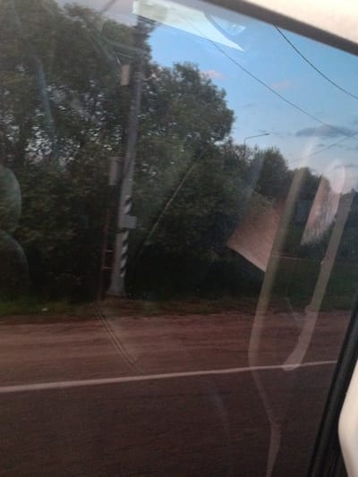 The image shows a view through a car window. Outside the window, there are trees, a utility pole, and a road. The sky is visible and appears to be partly cloudy. Reflections are visible on the car window, obscuring some of the details of the outside scene. The road appears to be paved and has a white line marking the lane. The scene suggests a rural or suburban setting, possibly during the daytime. There are no people visible in the image. The location is identified as Kardonikskaya, Russia, though there are no specific landmarks within the image to confirm this location.