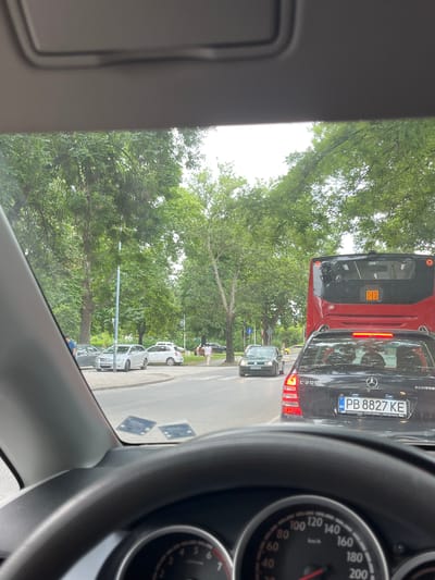 The image captures a traffic scene from the perspective of a car driver in Orizari, Bulgaria. In the foreground, the steering wheel and part of the dashboard are visible. Ahead, a black Mercedes-Benz C200 KOMPRESSOR is in traffic, with its license plate reading "PB 8827 KE". To the left of the Mercedes, a dark green Volkswagen Golf is also in traffic, with its license plate partially visible as "PB 5497 BM". Further ahead, a red bus is stopped in traffic. The surrounding environment features mature trees lining a paved road. Two small square stickers are visible on the interior of the car's windshield. The sky is overcast.