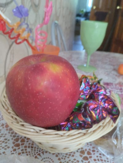 A close-up, slightly out-of-focus shot shows a red apple in a woven basket. The apple is the dominant subject, filling the left side of the frame. The basket contains colorful, individually wrapped candies with shiny foil wrappers in shades of purple, green, and gold. In the background, a pale green goblet is visible, along with other indistinct objects and a light-colored wall. The scene appears to be indoors, possibly on a table covered with a patterned tablecloth.