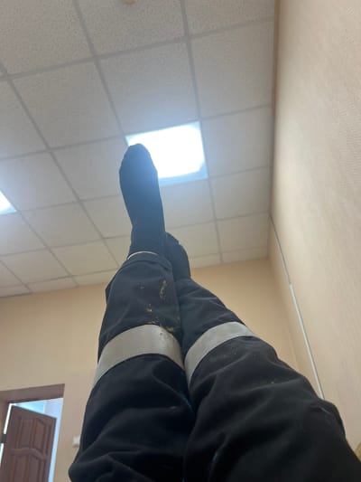 A low-angle indoor shot depicts a person lying down, their legs crossed at the knees. The person wears dark pants with reflective white stripes around the lower legs and black socks. The backdrop features a drop ceiling with recessed fluorescent lighting. Part of a tan wall and the top of a dark-colored door are visible in the lower right corner. The location is Бизяки, Russia.