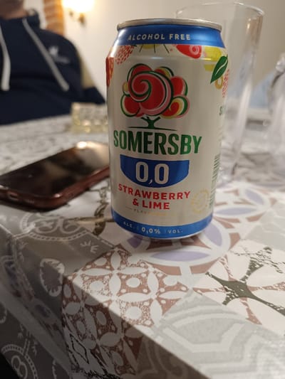 This image depicts an indoor scene in Izbeglii, Bulgaria, focused on a table surface. In the foreground, slightly right of center, stands an aluminum can of Somersby 0.0 Strawberry & Lime flavored non-alcoholic beverage. The can is primarily silver with a blue top and base band, displaying "ALCOHOL FREE" in blue lettering at the top, a stylized red and green tree logo, "SOMERSBY" in green, "0,0" in white on a blue oval, and "STRAWBERRY & LIME FLAVOURED" in red and green text. The can also states "ALC. | 0,0% | VOL." at its base.

To the left of the can, a dark-cased smartphone lies face down on the table. Behind the can, a clear, empty drinking glass is visible. The objects rest on a tablecloth featuring a repetitive pattern of light brown, white, and gray geometric and floral motifs.

In the blurred background, a person is partially visible, seated. This individual is wearing a dark blue hooded sweatshirt with white drawstrings and an Adidas logo on the right chest area. Behind the person, a light-colored wall (possibly brick) with a lit wall sconce or lamp is discernible, casting a warm glow. The overall scene suggests a casual, indoor setting.