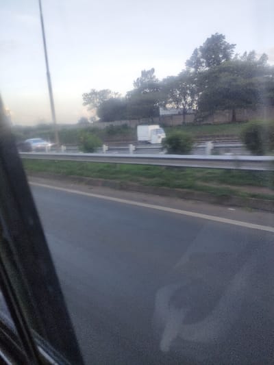 The scene depicts an exterior view from within a vehicle, looking out over a multi-lane road in Murera ward, Kenya. A dark, diagonal structure, likely a window frame, occupies the lower left quadrant. In the immediate foreground, an asphalt road surface with a solid white line delineates the lane or shoulder. Parallel to this, separated by a grassy median containing a silver metal guardrail, is another section of the road. On this parallel road section, a light-colored sedan with illuminated headlights is visible, traveling rightward. Further right on the same road section, a white three-wheeled light commercial vehicle, consistent with a Piaggio Ape, is also in motion. The background features a dense line of mature trees positioned above a light-colored wall or fence. The sky overhead is a uniform pale grey-blue, suggesting diffuse daylight. No human figures are discernible.