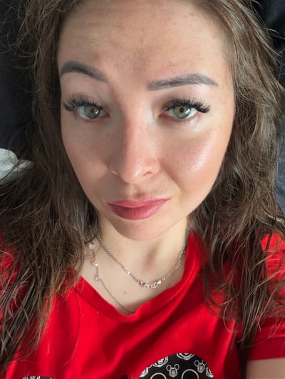 A close-up, overhead shot shows a woman with shoulder-length, dark brown, wavy hair and green eyes. She is wearing a bright red V-neck shirt with a silver necklace featuring small crystal pendants. The woman has light-colored skin with freckles, and appears to be wearing makeup, including mascara and lipstick. The background is dark.