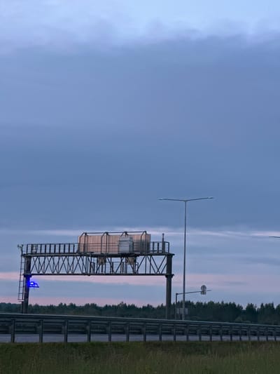 This is an image of a highway overpass with a blue light illuminated on a ladder leading up to it. The sky is overcast with shades of blue and grey, and there is a hint of pink from a sunset or sunrise. A line of trees forms a dark silhouette against the horizon. The overpass structure is made of metal beams and supports various pieces of equipment, possibly for traffic monitoring or toll collection. A street lamp is visible to the right of the overpass. The foreground shows a grassy verge and a metal guardrail separating the roadway from the shoulder. The location is identified as Budovo, Russia.