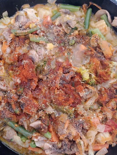 The image shows a close-up, overhead view of a dish being cooked in a dark pot. The dish appears to be a stew or curry, containing a variety of ingredients including green beans, carrots, mushrooms, corn, and pieces of meat. The food is seasoned with a generous amount of paprika, herbs, and black pepper. The overall impression is of a rustic, home-cooked meal. The image does not contain any specific details that would allow for its location to be identified as being in the city of Birsk, Russia, or any other city.