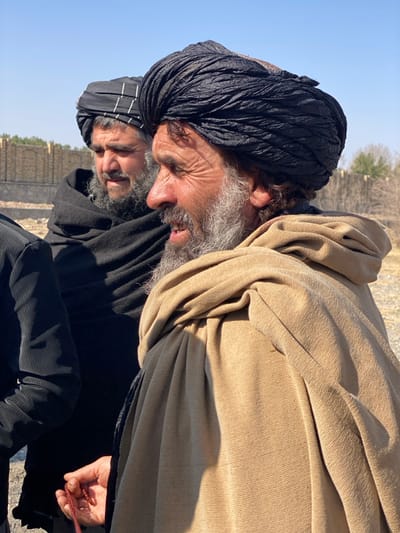 Two older men are in a landscape. One man has a black turban and grey beard. The second man has a black and white turban with a grey beard. The first man wears a tan shawl-like garment. The other man wears a black jacket. Trees and a crumbling wall are in the background. They both look down and to their right. One of the men holds a red beaded item.