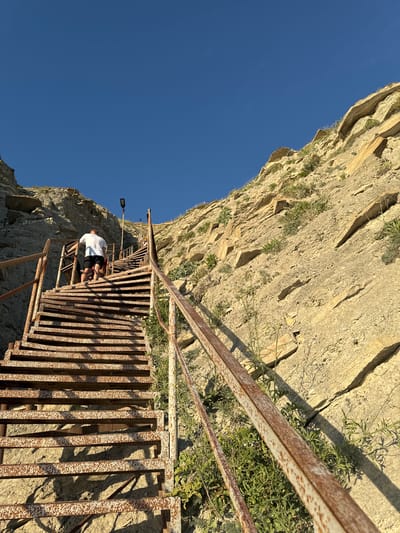 The image captures a steep, rusty metal staircase ascending a rugged, rocky cliff face under a clear blue sky. The staircase appears old and weathered, with visible rust and a slightly uneven structure, suggesting it might be a challenging but scenic route. Sparse green vegetation grows from cracks and crevices in the rock, adding a touch of nature to the otherwise stark landscape.
A person, wearing a white t-shirt and dark shorts, is seen ascending the stairs, appearing to be taking a break or looking around. Their posture suggests a moment of rest during the climb. The overall scene evokes a sense of adventure and physical exertion, with the immense scale of the cliff and the precariousness of the stairs contributing to the feeling of a remote or rugged location.
The lighting indicates it is daytime, likely late afternoon or early morning, given the long shadows cast by the stairs and the warm tone of the rock. The weather appears to be clear and sunny. The location is identified as Supsekh, Russia. There is no visible text in the image.
