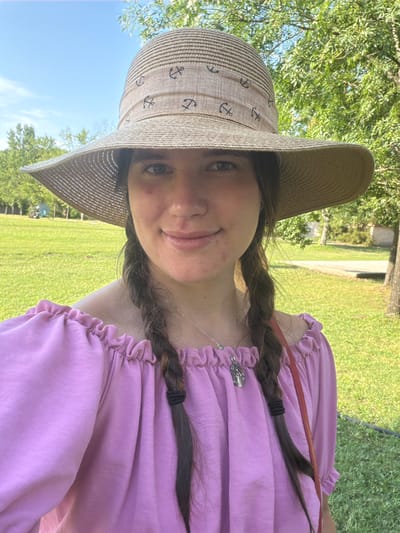A young woman is taking a selfie outdoors. She is wearing a wide-brimmed, straw hat with an anchor pattern and a ruffled, off-the-shoulder pink top. Her dark hair is in two braids. She is smiling slightly and has some blemishes on her cheek. The background shows a grassy area with trees and a blue sky. The scene appears to be a park or outdoor recreational area in Kiskunmajsa, Hungary.