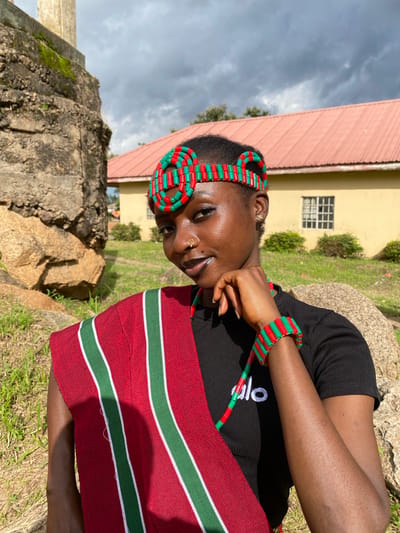 In Wang, Nigeria, a young Black woman, wearing a black t-shirt with the white word "alo" on it, poses for a photo outdoors. She has dark skin, dark hair, and is adorned with a red and green beaded headband and a matching bracelet. A red sash with green and white stripes is draped over her left shoulder. Her right hand rests on her chin, and she is wearing a small gold stud nose ring. The background includes a textured stone structure on the left, grassy terrain, and a yellow building with a red roof and a window on the right. The sky is cloudy.