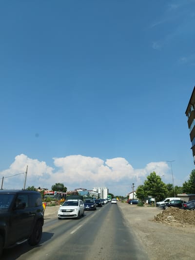 A vertical, eye-level shot depicts a road with moderate traffic in Bragadiru, Romania, under a bright blue sky with scattered white clouds. The scene focuses on the road and surrounding elements.
On the left side of the road, a black SUV is prominently featured, partially obscuring a white car driving in the same direction. Further ahead, a white hatchback with a license plate visible is in the left lane, followed by several other cars. The road appears to be asphalt, with visible lane markings.
To the right of the road, there is a dirt shoulder with some construction debris and parked vehicles, including a white van and a black sedan. Trees and buildings are visible in the background, suggesting an urban or semi-urban environment. A tall streetlight is positioned on the right side of the road. The overall impression is a typical day with traffic congestion in a Romanian town.