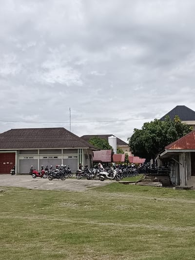 The image captures a cloudy day scene in Mesjid Reuntoh, Indonesia, featuring a parking lot filled with numerous motorcycles in front of several buildings. The foreground is dominated by a large, flat expanse of green grass, suggesting an outdoor public space or a park.
In the midground, a paved area serves as a parking lot. It is densely populated with a variety of motorcycles, most of which appear to be scooters or smaller commuter bikes. Many of the motorcycles have helmets on their handlebars or seats, indicating recent use. Several people, dressed in casual clothing, are visible among the motorcycles, some standing and others seemingly in the process of parking or preparing to leave.
Behind the parking lot, several structures are present. The most prominent building on the left has a long, dark tiled roof and a reddish-brown double door. It appears to be a functional building, possibly an office or a community center, with a row of rectangular windows along its facade. To the right of this building, a line of smaller structures with reddish-brown, corrugated metal roofs are visible, suggesting a more informal arrangement of buildings or covered areas. Further to the right, a larger building with a dark, angular roof is partially obscured by a large, leafy green tree. Another smaller, rustic structure with a rusted metal roof stands in the far right of the midground, adjacent to the larger building.
The sky is overcast with diffused grey clouds, creating a soft, even light across the scene. The overall impression is one of a busy but orderly outdoor environment on a day with moderate weather conditions.