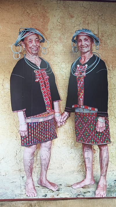 Two elderly women, wearing traditional attire, are depicted full-length in a photographic print, ostensibly located in Jiyang District, China. They stand barefoot, smiling directly forward, and are holding hands in the center. Both women wear black, short-sleeved tunics featuring central red, white, and black geometric patterns, and knee-length woven skirts with horizontal striped and cross-stitch motifs in red, green, black, and white. Their wrists are adorned with multiple silver-colored bangles, and their necks with multi-strand silver-colored necklaces. Elaborate headpieces, constructed from multiple silver-colored wire rings and large side hoops, crown their heads. Facial markings or tattoos are visible around both women's mouths and cheeks. Their exposed lower legs display distinct geometric tattoos; the left woman exhibits angular lines, while the right woman's legs feature several 'L'-shaped designs. They are positioned against a textured, cracked, yellowish-brown background.
