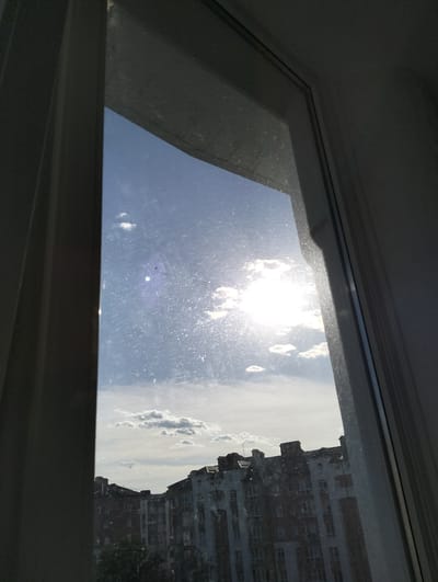 The image is taken from inside a building, looking out of a dirty window. The view is of a bright, sunny sky with scattered clouds. Below the clouds, a large, multi-story building complex is visible. The building's architecture consists of several sections of varying heights and designs, mostly a mixture of white and red brick. The structures contain numerous windows, suggesting residential apartments. Some trees are visible at ground level, partially obscuring the base of the building. Based on the description the image contains buildings found in the city of Krykhivtsi, Ukraine.