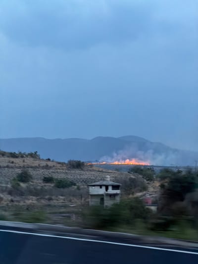 The image shows a landscape in the city of Santa Cruz Tehuixpango, Mexico, likely taken from a moving vehicle on a road. A significant wildfire is visible in the distance, emitting smoke and orange flames. In the foreground, there is a rocky, mountainous terrain with sparse vegetation, including a partially visible structure that appears to be a building. Further back, there is a mountain range silhouetted against the sky. The sky is overcast and blue. The right edge of the image shows part of a vehicle window.