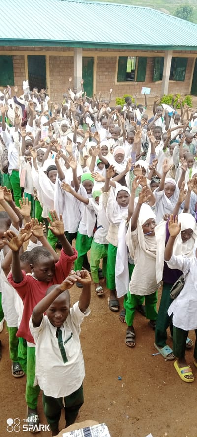 In Kagarko North, Nigeria, a large group of approximately 100-150 school-aged children, predominantly boys with some girls visible, are gathered outdoors in front of a building. The building appears to be a single-story structure with a green corrugated metal roof and a brick facade. The children are mostly wearing white or light-colored garments, many with green accents, and some girls are wearing hijabs. The majority of the children have their arms raised in the air, suggesting engagement in an activity such as a lesson, a song, or a greeting. The ground is a dirt or sandy surface. The overall scene conveys a sense of collective energy and participation.