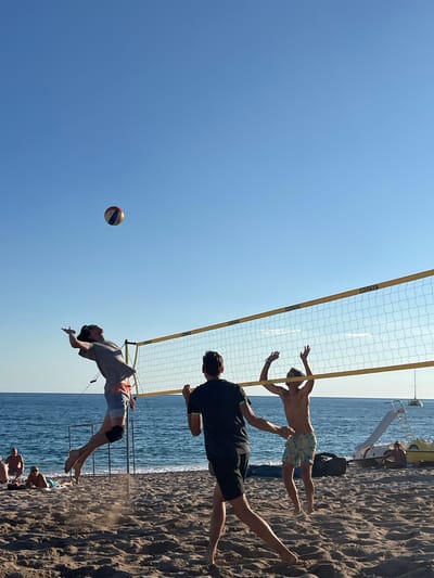 A group of young men are playing beach volleyball on a sandy beach next to the sea. The sky is clear and blue. A volleyball net is set up, and one man is jumping to spike the ball while another prepares to receive it. Two other men are also visible near the net, one in the foreground and one in the background. Several other people are sitting and relaxing on the beach in the background. A few beach chairs, umbrellas, and some equipment are scattered on the sand. The water appears calm, and a few sailboats are visible in the distance. The overall scene depicts a lively recreational activity on a sunny day at the beach.