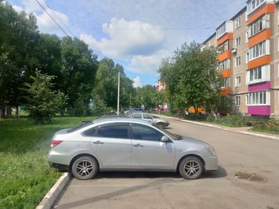 This is an exterior, medium-angle, eye-level, full-shot photograph taken on a cloudy day. In the foreground, a silver sedan is parked on a paved street, with its front right tire resting on the curb. The car's body shows scratches on the driver's side door. Behind the silver car, another silver sedan is parked, and further back, a light-colored sedan is visible. The street is lined with green grass and trees on the left and a multi-story apartment building with orange and pink accents on the right. The apartment building has numerous windows and balconies. A power line stretches across the upper portion of the image. The scene appears to be in a residential area, likely in the city of Salavat, Russia, given the architectural style of the building and the presence of typical urban vegetation. There are no people visible in the immediate foreground or mid-ground of the image.