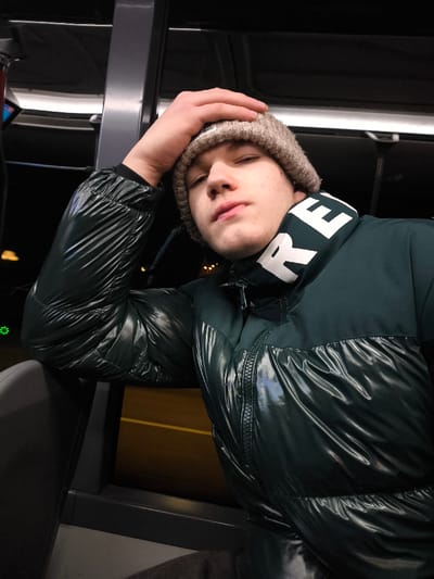 A young man with a light complexion is seated on public transportation. He wears a tan beanie and a shiny, dark green puffer jacket with a white collar featuring bold black lettering. His right hand rests on his head, and he leans against a dark grey partition. A blurry, illuminated window is visible behind him, suggesting nighttime. The scene is dimly lit, highlighting the jacket's glossy texture.