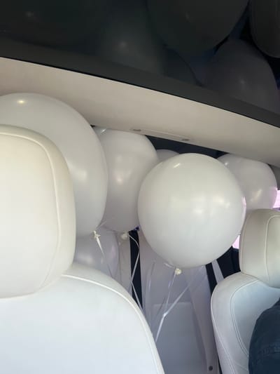 The media file presents a close-up view of a vehicle's interior, likely a car or van, completely filled with numerous white, glossy, inflated balloons. The balloons vary slightly in size, with light reflecting off their smooth surfaces. Many of them have thin white ribbons attached, which trail downwards.
Interspersed among the balloons are elements of the vehicle's interior. Sections of white upholstered seating, possibly car seats, are visible, characterized by their soft texture and subtle stitching. A prominent dark, reflective surface, likely a window or a dark panel of the vehicle's interior, frames the left side of the image, providing a strong contrast to the bright white objects. In the upper right corner, a small portion of dark fabric, resembling clothing, is visible, hinting at the presence of a person, though no individual is clearly depicted.
The scene suggests a celebratory event or a surprise, where a vehicle has been decoratively filled with balloons. The overall mood conveyed is one of joy and festivity. The image originates from Yeghvard, Armenia. No text is visible.