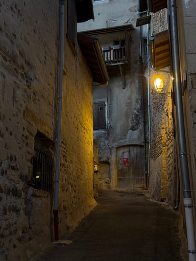 This image depicts a narrow alleyway in Saint-Antoine-l'Abbaye, France, at dusk or night. The scene is lit by a single, old-fashioned street lamp emitting a warm, yellow glow, casting long shadows on the cobblestone path and ancient stone walls. The buildings on either side of the alley are constructed from rough-hewn stones and appear aged, with visible signs of wear and tear. One building features a wooden door with a small, red sign attached. Above the door, a small, weathered balcony with potted plants is visible. A drainpipe runs down the side of one building, parallel to the alley. The overall atmosphere is one of quiet solitude and historical charm. There are no people or animals visible in the image.