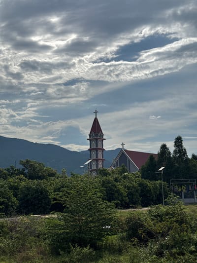 The image depicts a scenic view in Suối Tân, Vietnam. The main subjects are a church tower with a red roof and cross, lush green trees, and a mountain range in the background. The sky is filled with dramatic, wispy clouds, suggesting a dynamic weather pattern, possibly late afternoon or early evening. The overall mood is serene and picturesque.
The church tower, visible behind a dense canopy of trees, features intricate architectural details. Its pointed roof is a vibrant red, contrasting with the muted tones of the surrounding landscape. A cross stands prominently at its apex. The trees are in full foliage, indicating a warm season. The mountain range appears to be covered in a dark, possibly forested, terrain.
The setting seems to be a rural or semi-rural area, given the presence of the church and the natural landscape. There are no people visible in the frame, but a paved pathway is partially obscured by the trees on the right side of the image. A street lamp is also visible among the trees.
The orientation of the image is vertical, rotated 90 degrees clockwise. The lighting suggests it is daytime, with the sun casting light on the clouds and the church. The clouds are a significant feature, adding depth and texture to the sky.
There is no visible text in the image.